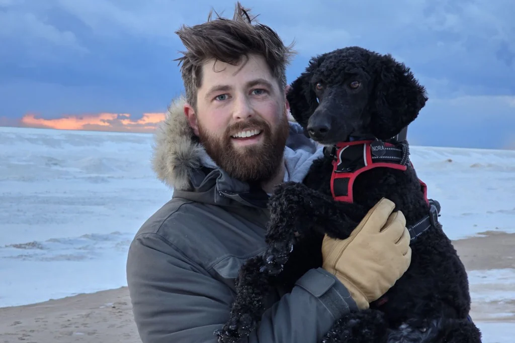 Joe Despres holding a black dog on a snowy beach at sunset